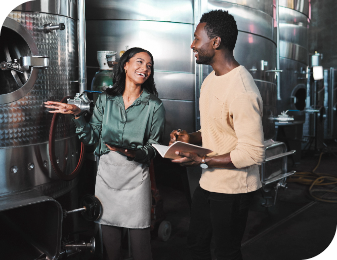 Two people, a woman and a man, are happily talking in a brewery or industrial setting. The woman is holding a tablet and gesturing towards large stainless steel tanks. She is dressed in a green shirt that makes her look professional, yet practical. The man is taking notes in a notebook.