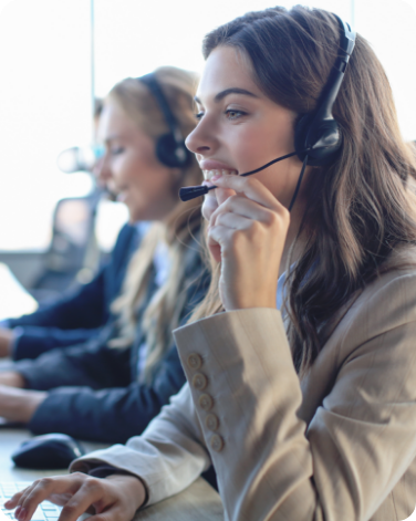 Two women with headsets, likely customer service representatives, working in a call center. The brunette woman in the foreground, who is in focus, has a thoughtful expression and is holding the microphone part of her headset. The blond woman in the background is out of focus. Both are wearing professional attire and are positioned in front of computers.