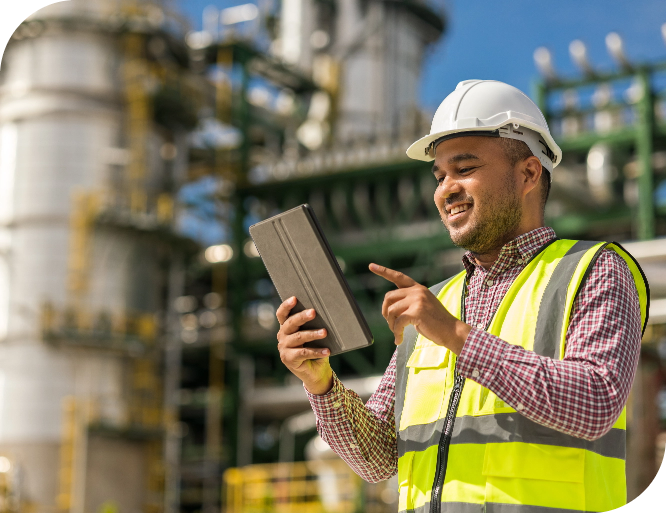 An engineer wearing a security helmet and high-visibility vest is smiling and holding a tablet, in front of an outdoors industrial facility with pipes and steel structures in the background.