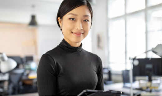 An asian woman is standing in an office environment, smiling at the camera. She is wearing a black turtleneck and holding a tablet. The background is softly out of focus.