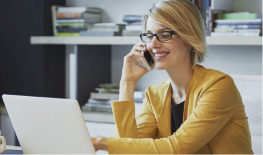 Short general shot of a professional woman in an office environment. She's blonde, wearing glasses, a yellow blazer, and is smiling while talking on a smartphone with one hand and using a laptop with the other.