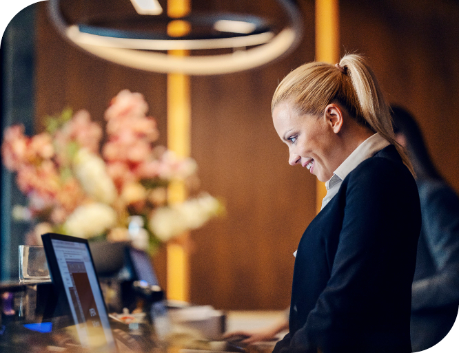 A woman with blonde hair tied back, wearing a dark blazer and a white shirt, is working at a reception desk at a lobby. She’s smiling and focused on a computer screen. The background features warm ambient lighting and blurred floral arrangements.